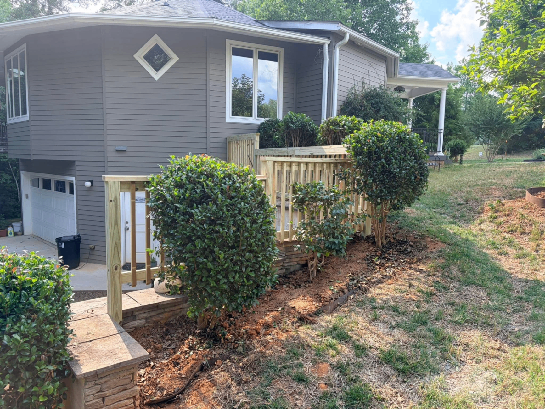 Side entry deck with new pine railings and stacked-stone steps beside a gray home